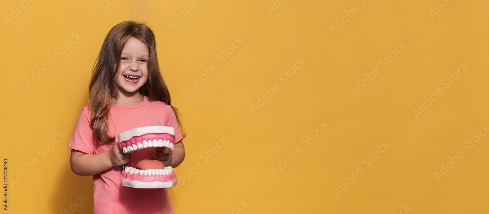 Wall mural A smiling girl with healthy teeth holds a large jaw in her hands on a yellow isolated background. A place for your text. Oral hygiene. Pediatric dentistry. Prosthetics. Rules for brushing teeth.