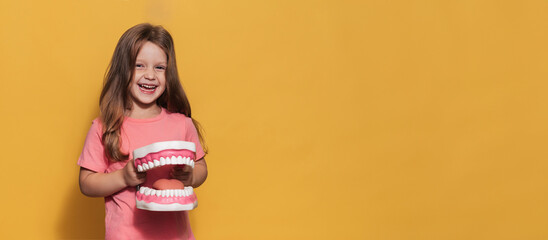 A smiling girl with healthy teeth holds a large jaw in her hands on a yellow isolated background. A place for your text. Oral hygiene. Pediatric dentistry. Prosthetics. Rules for brushing teeth.