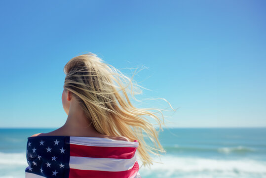 AI Generated Image. Young Blond woman with long hair wrapped in American US flag looking at the ocean