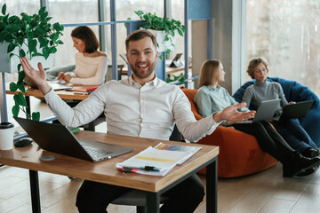 Happy man is smiling, sitting by table. People are working in the office with bean bags chairs in it