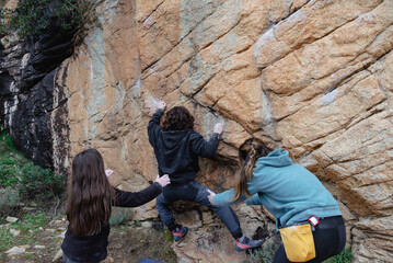A climber is carefully ascending a steep rock face, guided and supported by two companions, against a backdrop of natural mountain terrain
