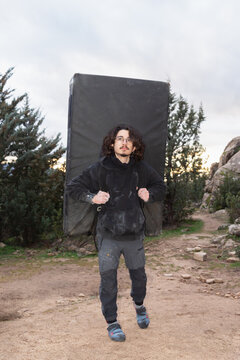 A focused boulderer stands with a large crash pad on his back, ready to embark on an outdoor climbing adventure in a rugged mountain setting