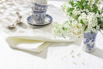 Spring tea party serving. Morning bright light at the garden, white tablecloth and cups, flowers. Fresh bright atmosphere