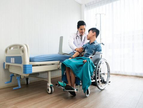Compassionate Asian pediatric doctor listen and use measure pressure to a young handicapped child patient sitting on wheelchair beside a hospital bed with caring atmosphere in recovery room