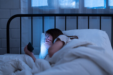 Asian woman lies in bed late at night while looking at her phone gadget device with a worried...