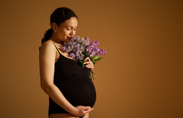 Beautiful pregnant woman in black bodysuit, holding her big belly while posing with a bouquet of...