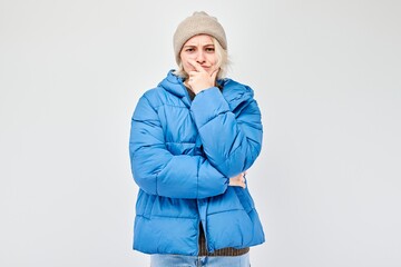 Woman in blue winter jacket and beanie with a thoughtful expression, isolated on a light background.