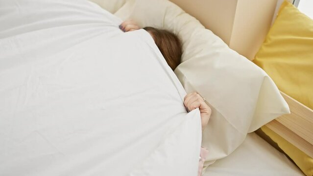 A woman hides under a white duvet in a bright, cozy bedroom setting, evoking a sense of morning routines.