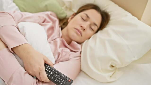 Young woman asleep in bed with a remote control in her hand, depicting relaxation in a comfortable home environment.
