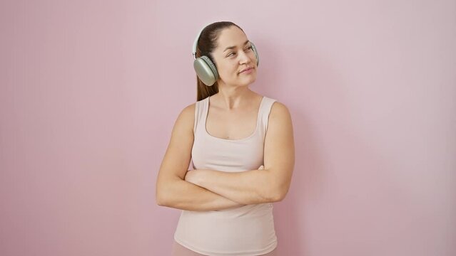 Cheery young woman in sportswear, jamming to music on headphones. her blue eyes sparkling, she smiles, looking away, lost in positive thoughts against a pink isolated background.
