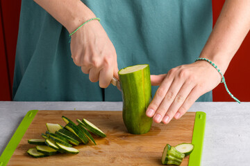 Authentic female hands cutting zucchini on wooden cutting board on kitchen table. Woman in apron...