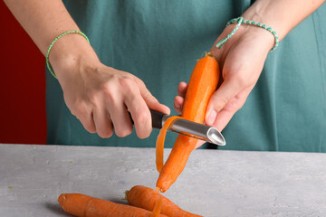 Authentic female hands peeling fresh carrot at wooden table with a stainless knife at kitchen table, close up, vegan and vegetarian food lifestyle, domestic life, preparing food