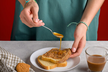Woman's hands puting honey with a spoon on a toast with peanut butter to make a sandwich for breakfast, at gray kitchen table, close-up. Typical snack food, food lifestyle, domestic life