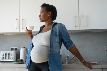 Pregnant woman holding tea cup in kitchen - obrazy, fototapety, plakaty