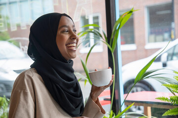 Smiling young woman in hijab drinking coffee in cafe - Powered by Adobe