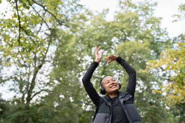 Portrait of smiling woman with headphones raising arms in park