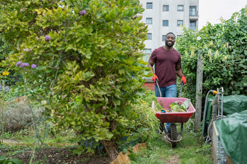 Portrait of smiling man with vegetables in wheelbarrow in urban garden