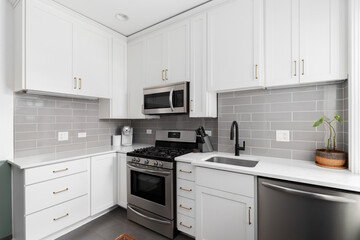 A small, white kitchen with a grey subway tile backsplash, stainless steel appliances, and grey...