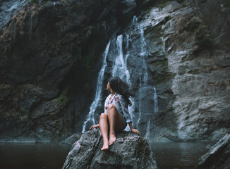 Portrait of a young woman sitting on a log and looking at a waterfall. - Powered by Adobe