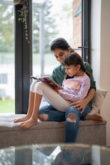 Mother and daughter sitting near window and reading book