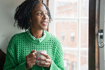 Smiling woman holding cup of coffee while looking through window © Cultura Creative