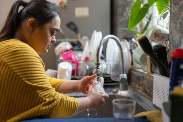 Young woman with down syndrome washing dishes at home - obrazy, fototapety, plakaty