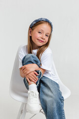 Stylish little girl wearing white t-shirt and jeans looking at camera sitting on studio white background