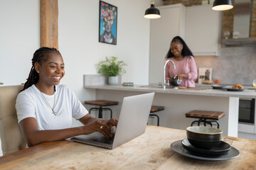 Woman working on laptop while girlfriend preparing breakfast in kitchen - obrazy, fototapety, plakaty