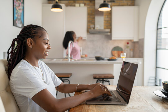 Woman working on laptop while girlfriend preparing breakfast in kitchen