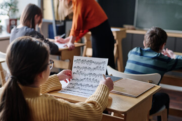 Selective focus shot of unrecognizable girl sitting at desk in classroom reading sheet music during class at school