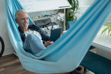 Smiling mature man lying in hammock and using digital tablet - obrazy, fototapety, plakaty