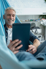 Smiling mature man lying in hammock and using digital tablet