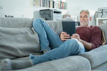 Smiling mature man using digital tablet on sofa - obrazy, fototapety, plakaty