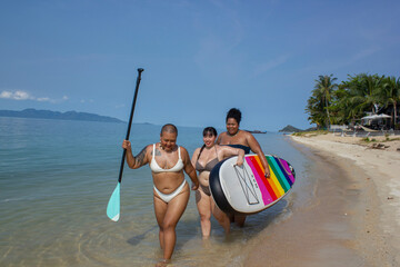 Group of cheerful women walking with paddleboard along beach