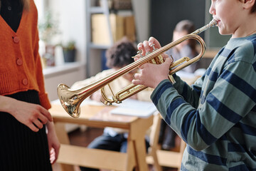Selective focus crop shot of Caucasian boy standing in front of teacher playing trumpet during...