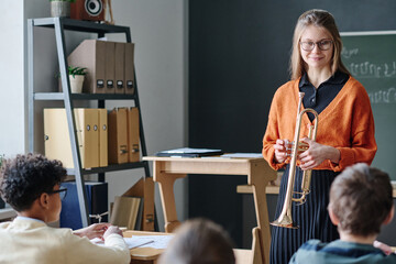 Young female music teacher holding trumpet standing in classroom in front of kids conducting class