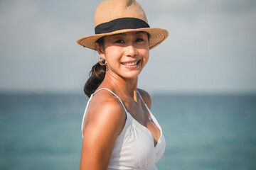 Portrait of smiling young woman wearing a straw hat standing at the beach. Asian woman on vacation...