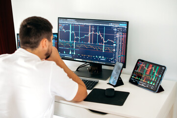 Stock trading, cryptocurrency concept. Concentrated male trader sitting at desk at home office monitoring stock market looking at monitors analyzing price flow, risks and prospects