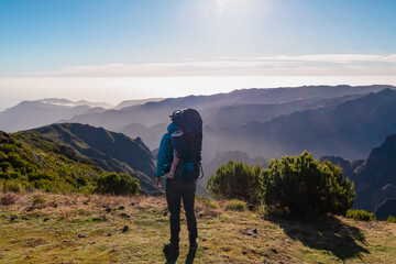Hiker man with baby carrier backpack looking at misty hills and canyon of rugged terrain on Madeira island, Portugal, Europe. Idyllic hiking trail to mountain peak Pico Ruivo. Remote coastal landscape