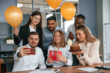 Making selfie with red colored smartphone, with cake. Employee having a birthday in the office, group of workers