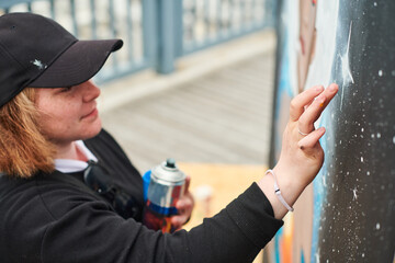 Female artist in black cap is painting picture with paint spray can spraying it onto canvas at...