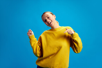 Positive, beautiful, smiling young girl in yellow sweatshirt dancing, having fun against blue...