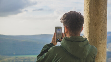 White man taking a picture with his phone wearing a green jacket and taking a landscape vertical picture of the countryside of Motovun, in the region of Istria in Croatia - Powered by Adobe