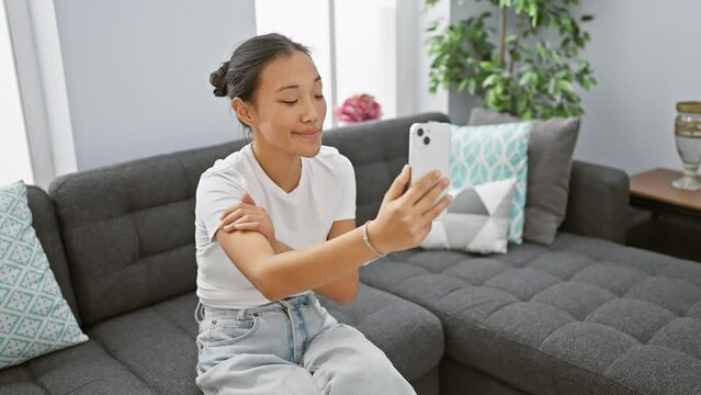 Asian woman video calling at home, showing discomfort in shoulder while sitting on couch.
