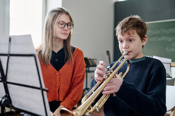 Medium shot of Caucasian schoolboy playing trumpet at music class, his female teacher listening to...