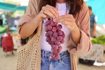 Cropped shot of a young woman with a net bag picking fresh grapes on farmers market. Shopping for...