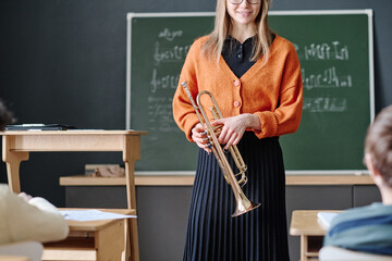 Crop shot of cheerful young female music teacher holding trumpet working with kids at school