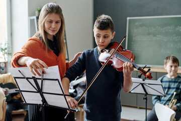 Young female music teacher working with student playing violin at music class at school