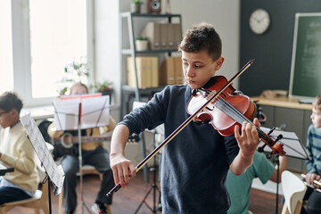 Medium shot of Caucasian boy practicing violin for school orchestra in classroom, his classmates...