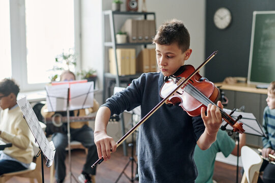 Medium shot of Caucasian boy practicing violin for school orchestra in classroom, his classmates sitting in background